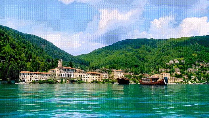 Scenic view of a calm lake surrounded by lush green mountains, featuring a quaint town with traditional buildings and several boats docked at the shore.